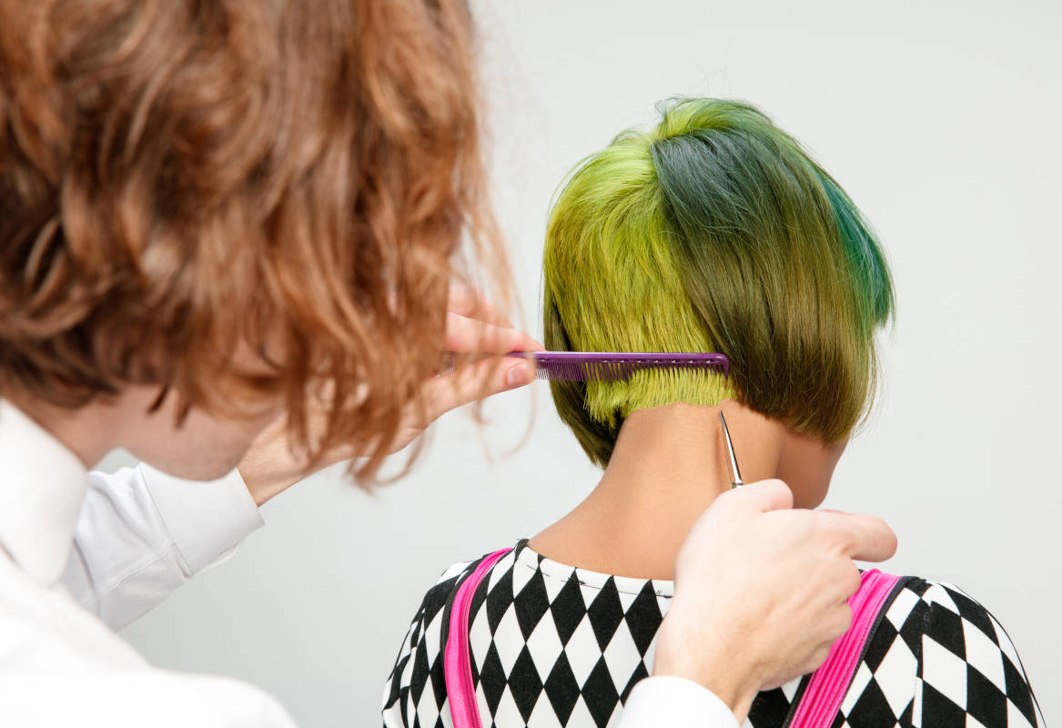 Picture showing adult woman at the hair salon. Studio shot of graceful young girl with stylish short haircut and colorful hair on gray background and hands of hairdresser.